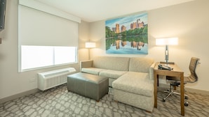 Une chambre d’hôtel avec une grande fenêtre, un canapé, un bureau avec une chaise, un radiateur et un tableau représentant un lac.
