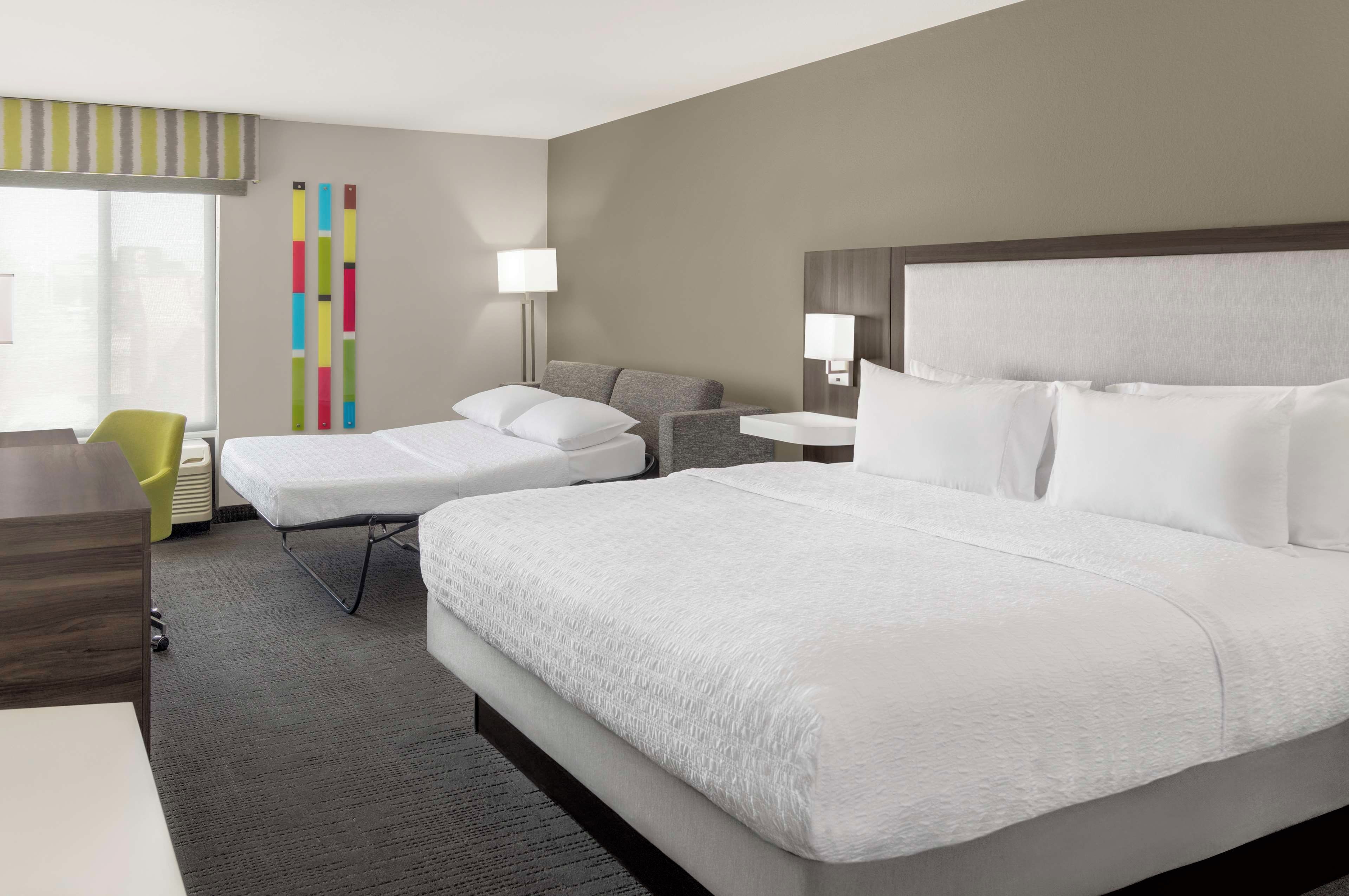 A hotel room with a large bed, a smaller bed, a desk, a chair, and a window with blinds.