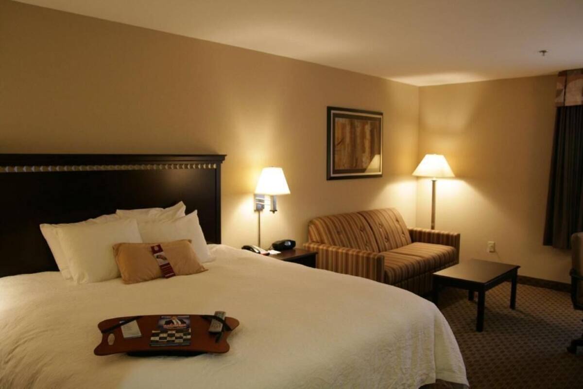Quarto de hotel com uma cama grande, duas luminárias de cabeceira, uma cadeira, uma mesinha e um quadro na parede.