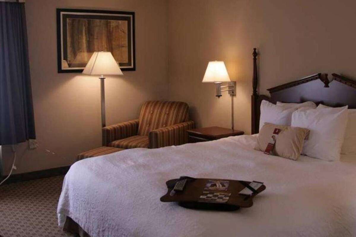 Quarto de hotel com uma cama, uma cadeira, mesa de cabeceira, abajur e um porta-bandeja com objetos.