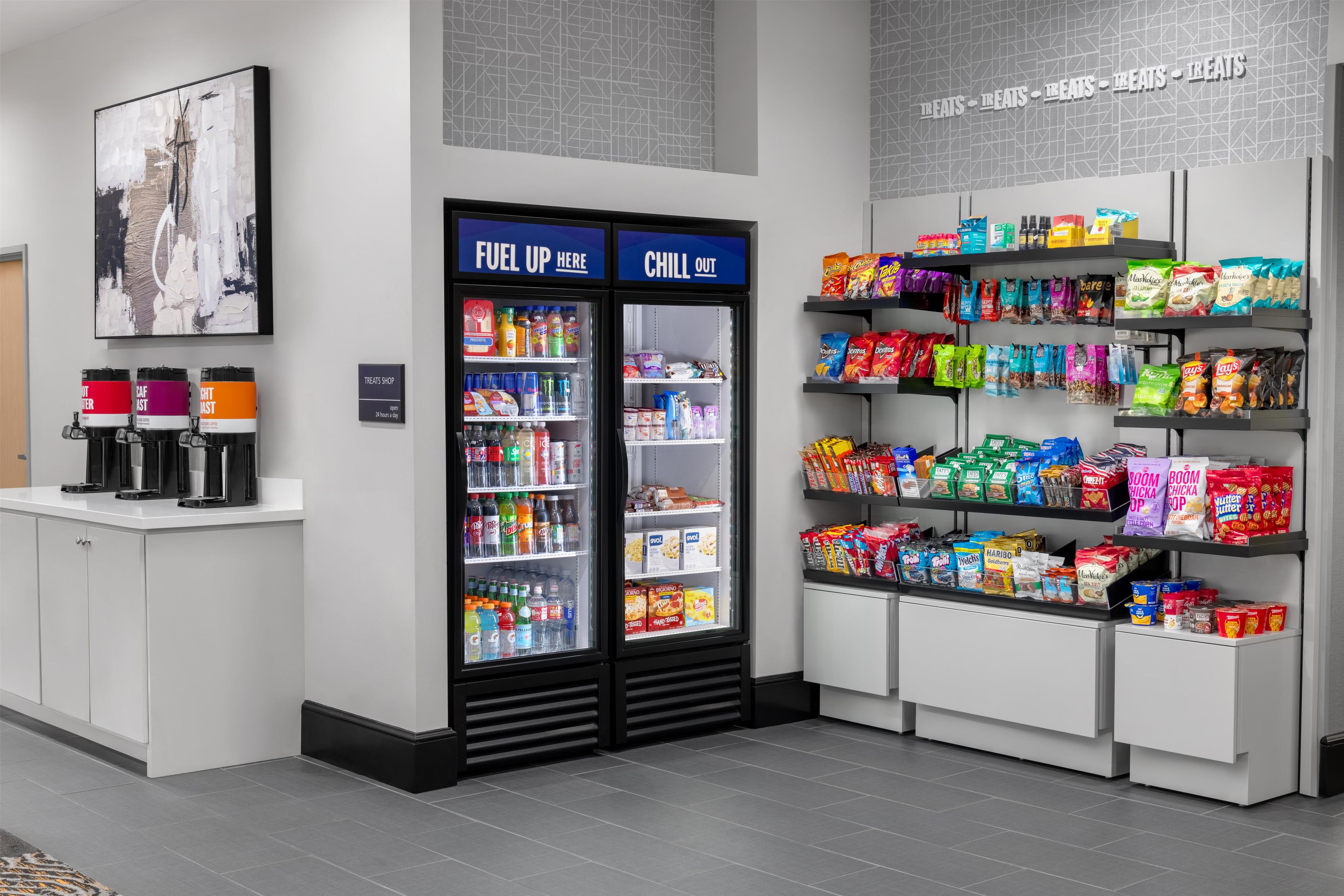 Un bar de aperitivos con un refrigerador para bebidas, estantes con bocadillos y una estación de café.