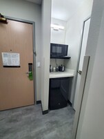 Un pasillo estrecho con una puerta a la izquierda, un microondas en un estante superior y una pequeña zona de kitchenette.