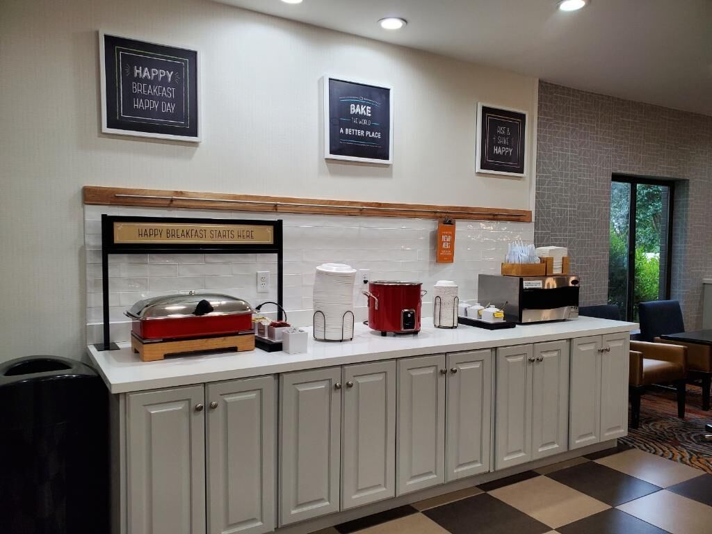 A breakfast bar with a sign that reads "HAPPY BREAKFAST STARTS HERE" and a red slow cooker. There are white plates stacked on the counter, a coffee maker, and a kettle. The wall has framed signs with messages like "HAPPY BREAKFAST HAPPY DAY" and "BAKE THE WORLD A BETTER PLACE."