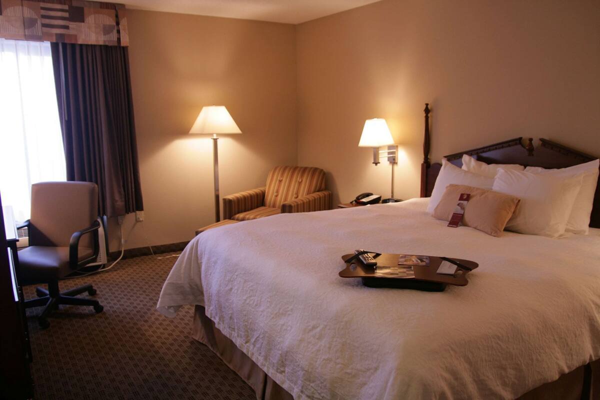 Quarto de hotel com uma cama grande, duas luminárias de cabeceira, uma cadeira, uma escrivaninha com um computador e um tabuleiro com objetos sobre a cama.