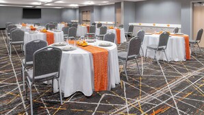 Un salón de conferencias con varias mesas dispuestas en filas, cada una con mantel blanco y camino de mesa naranja, y sillas grises.