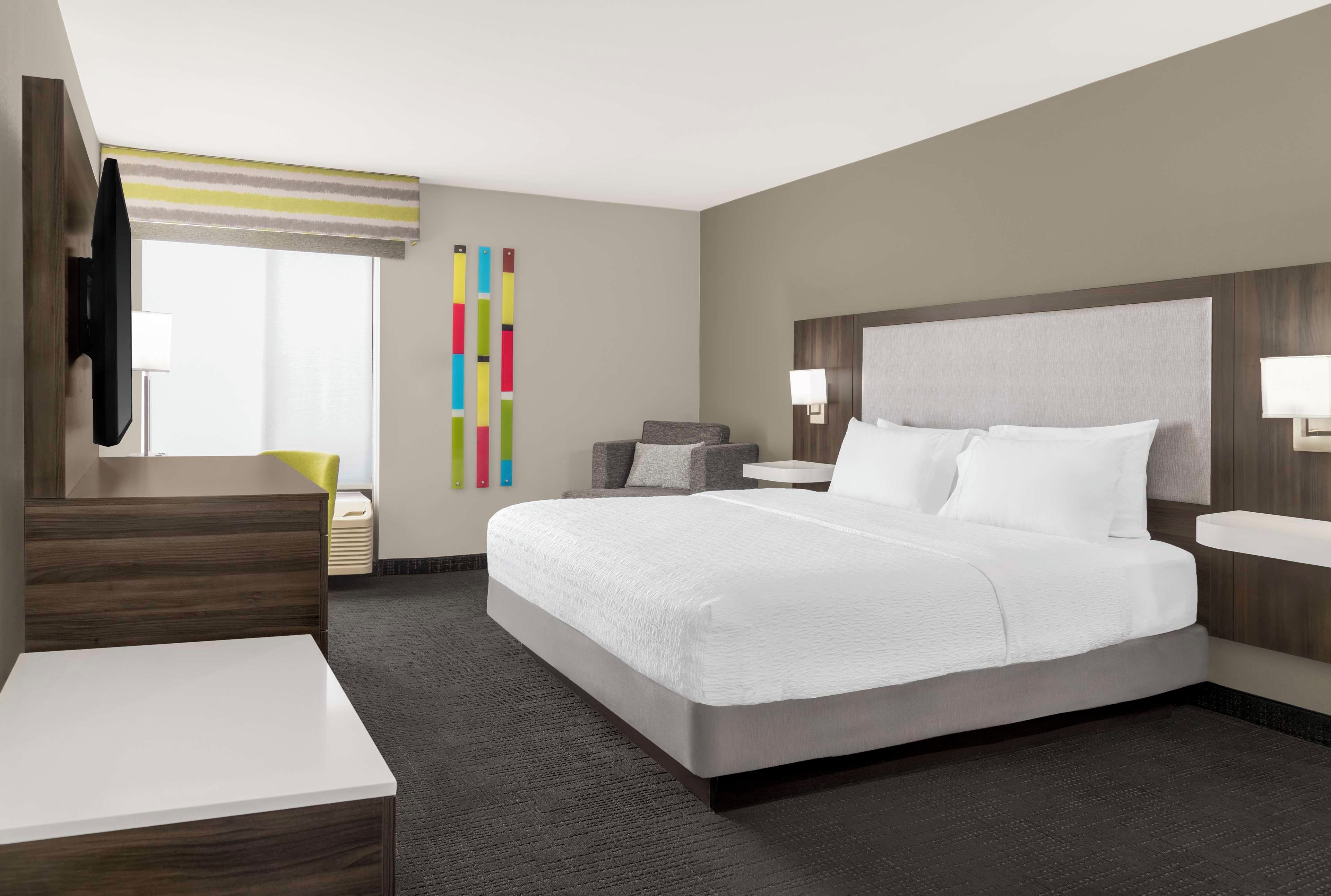 A hotel room with a large bed, a desk, a chair, a bedside table, a lamp, and a window with a view.