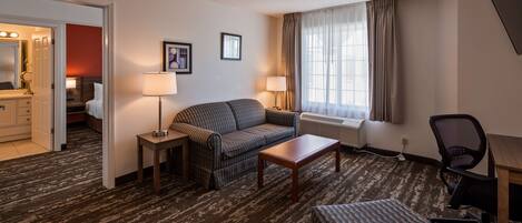A hotel room with a sofa, a small table, a chair, a lamp, a radiator, and a window with curtains.