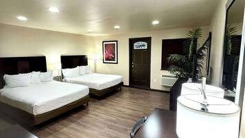 A hotel room with two beds, a desk, a lamp, a door, and a plant.
