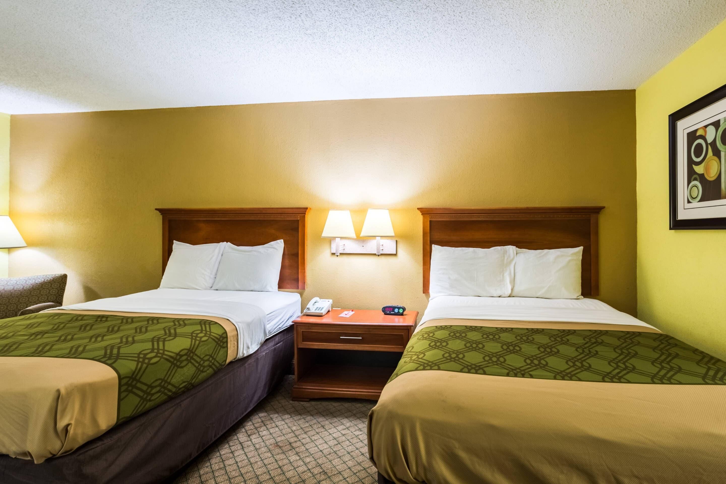 A hotel room with two beds, a nightstand with a phone and alarm clock, and a wall-mounted lamp.