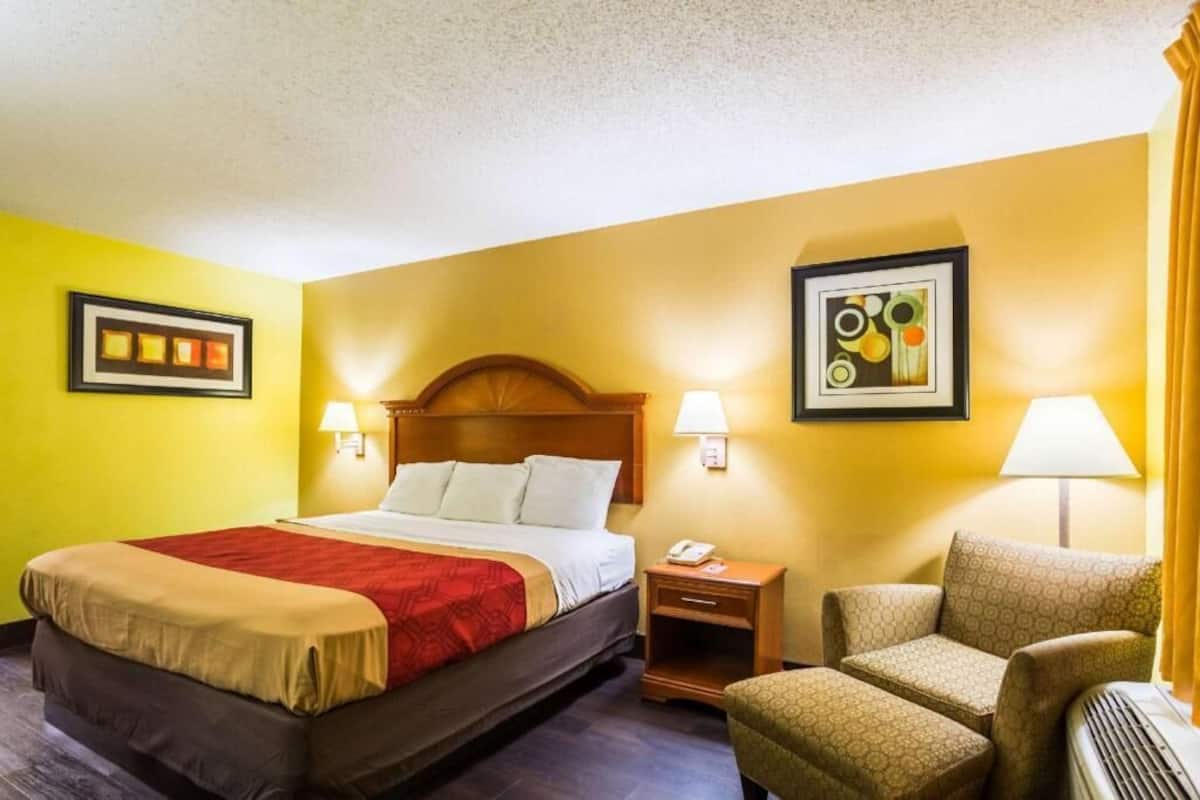 Habitación de hotel con cama, lámparas de noche, una silla, mesita de noche y un cuadro colgado en la pared.