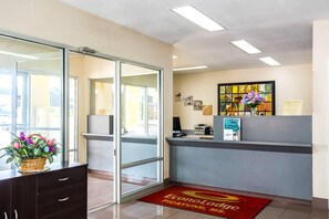 Zona de recepción con puerta de vidrio, un mostrador con computadora y una alfombra roja con un logotipo.