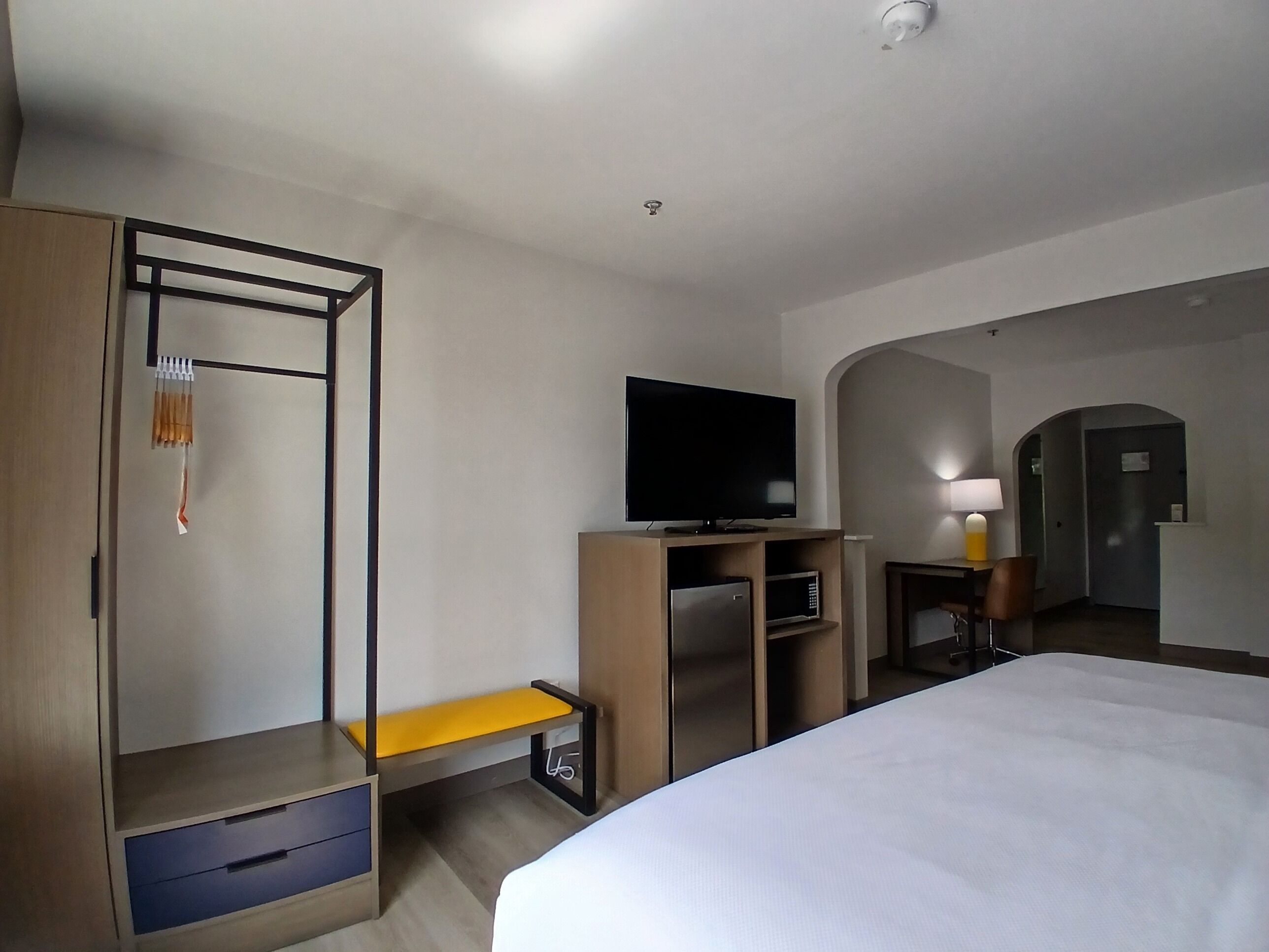 Habitación de hotel con cama, televisión, escritorio con silla, armario y una mesita de noche con lámpara.