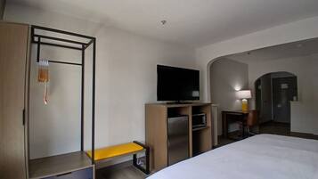 Habitación de hotel con cama, televisión, escritorio con silla, armario y una mesita de noche con lámpara.