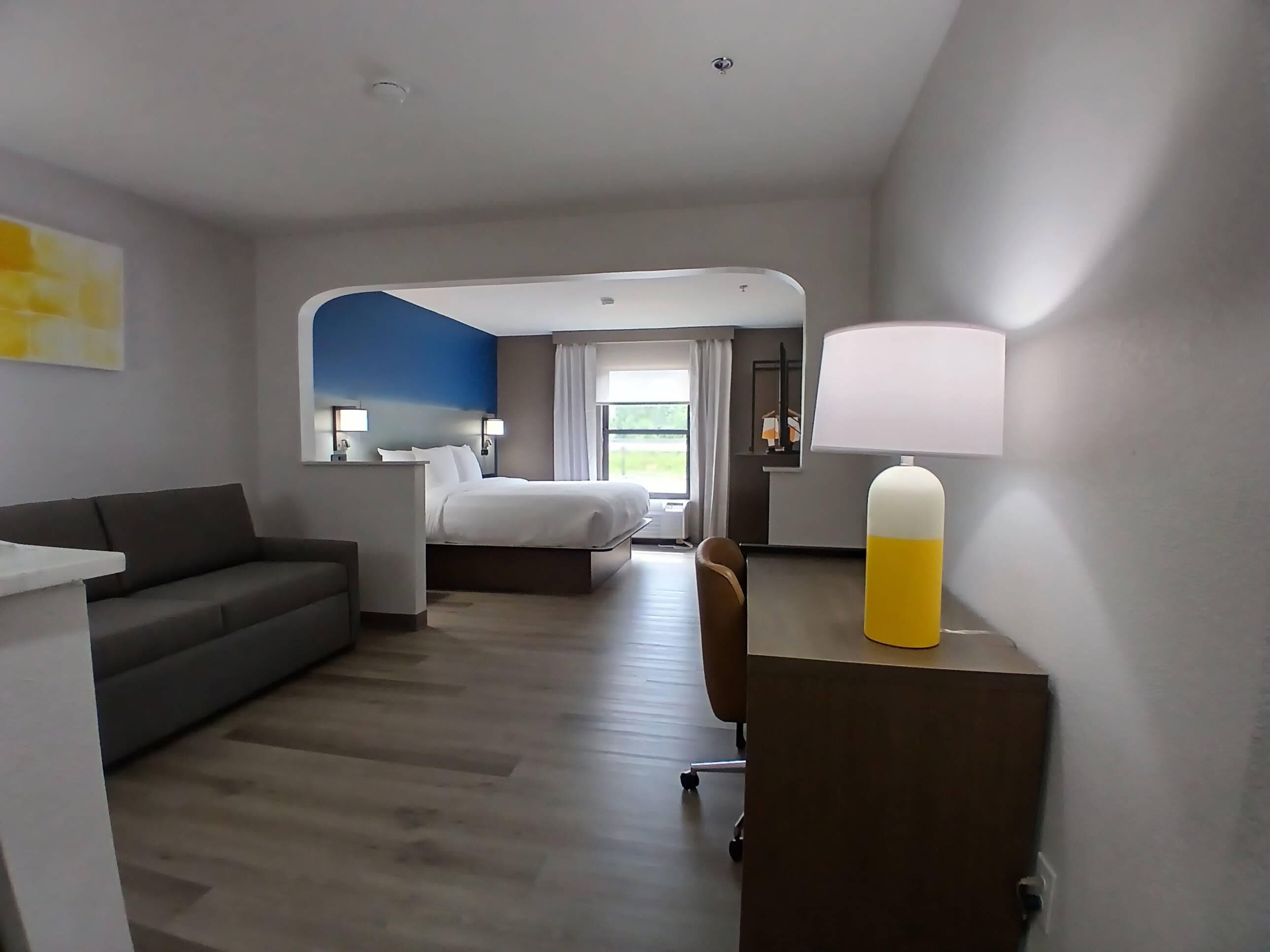 A modern hotel room with a bed, a desk, a chair, a sofa, and a lamp.