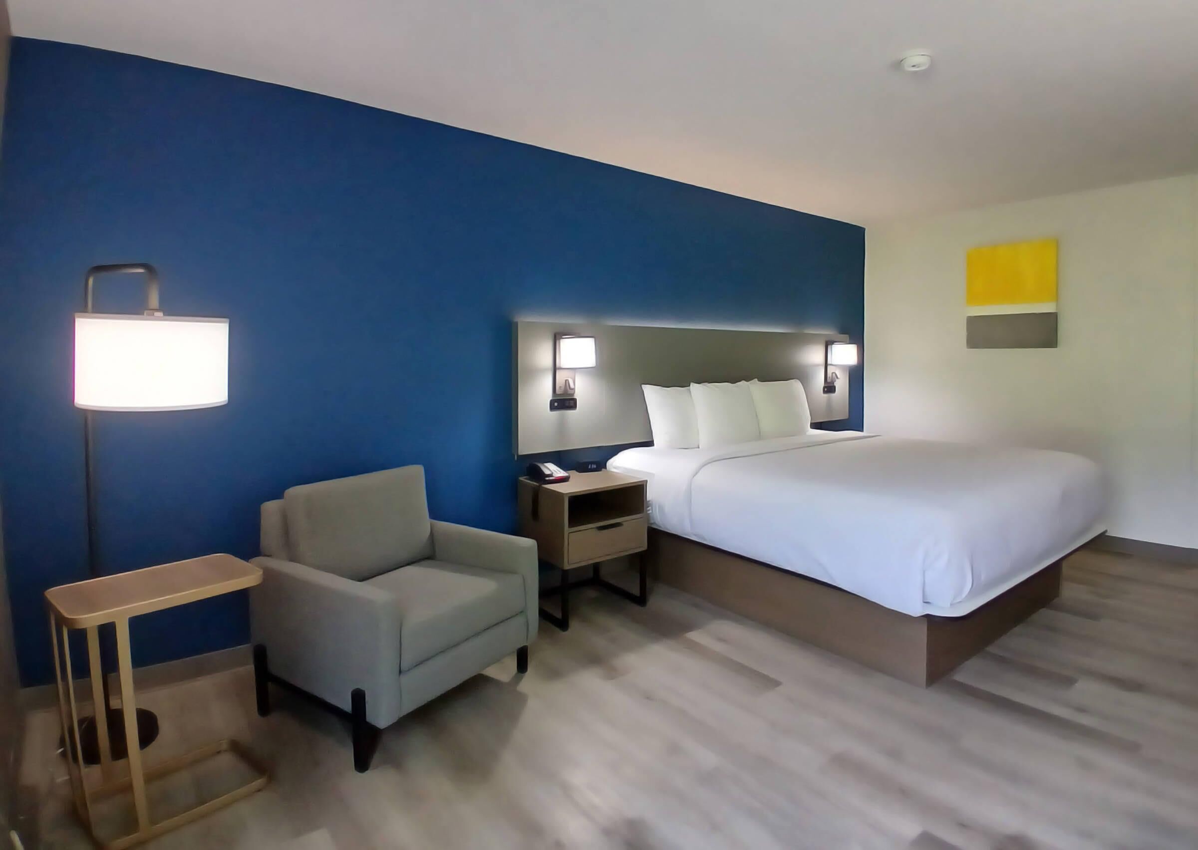 A hotel room with a bed, a chair, a nightstand, a lamp, and a wall with a light switch.