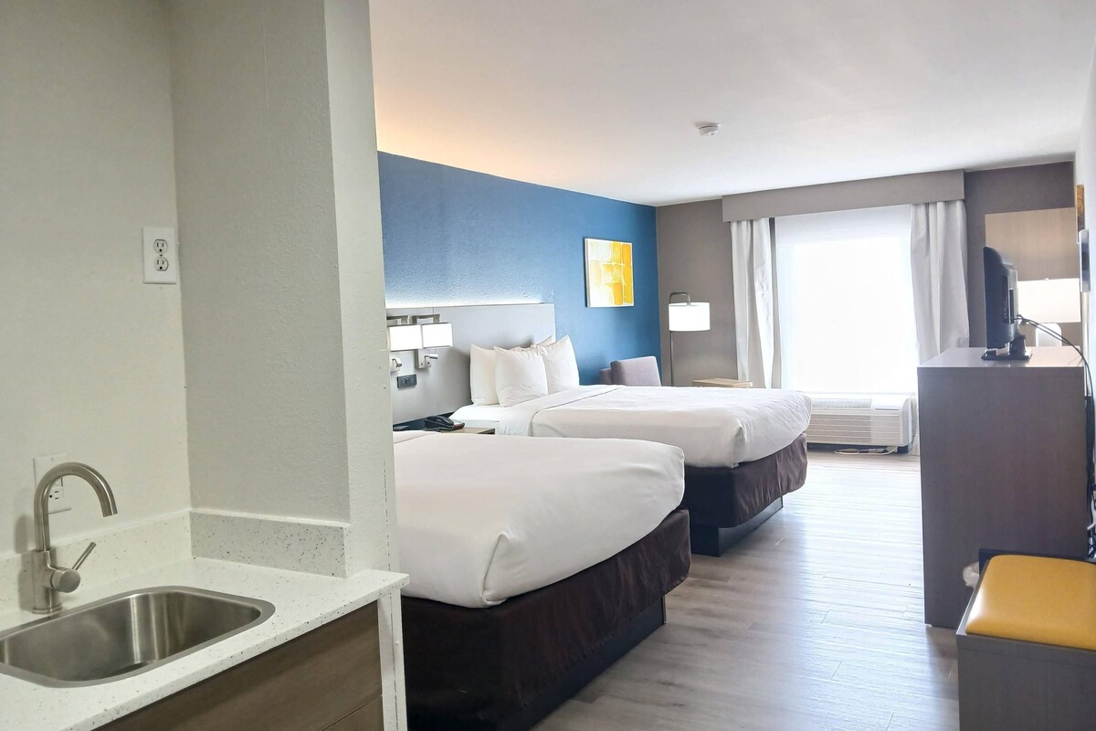 Ett hotellrum med en dubbelsäng, en enkelsäng, ett skrivbord med en stol, ett handfat och ett fönster med gardiner.