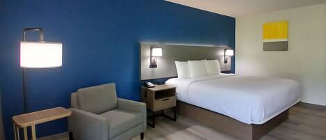 A hotel room with a bed, a chair, a nightstand, a lamp, and a wall with a light switch.