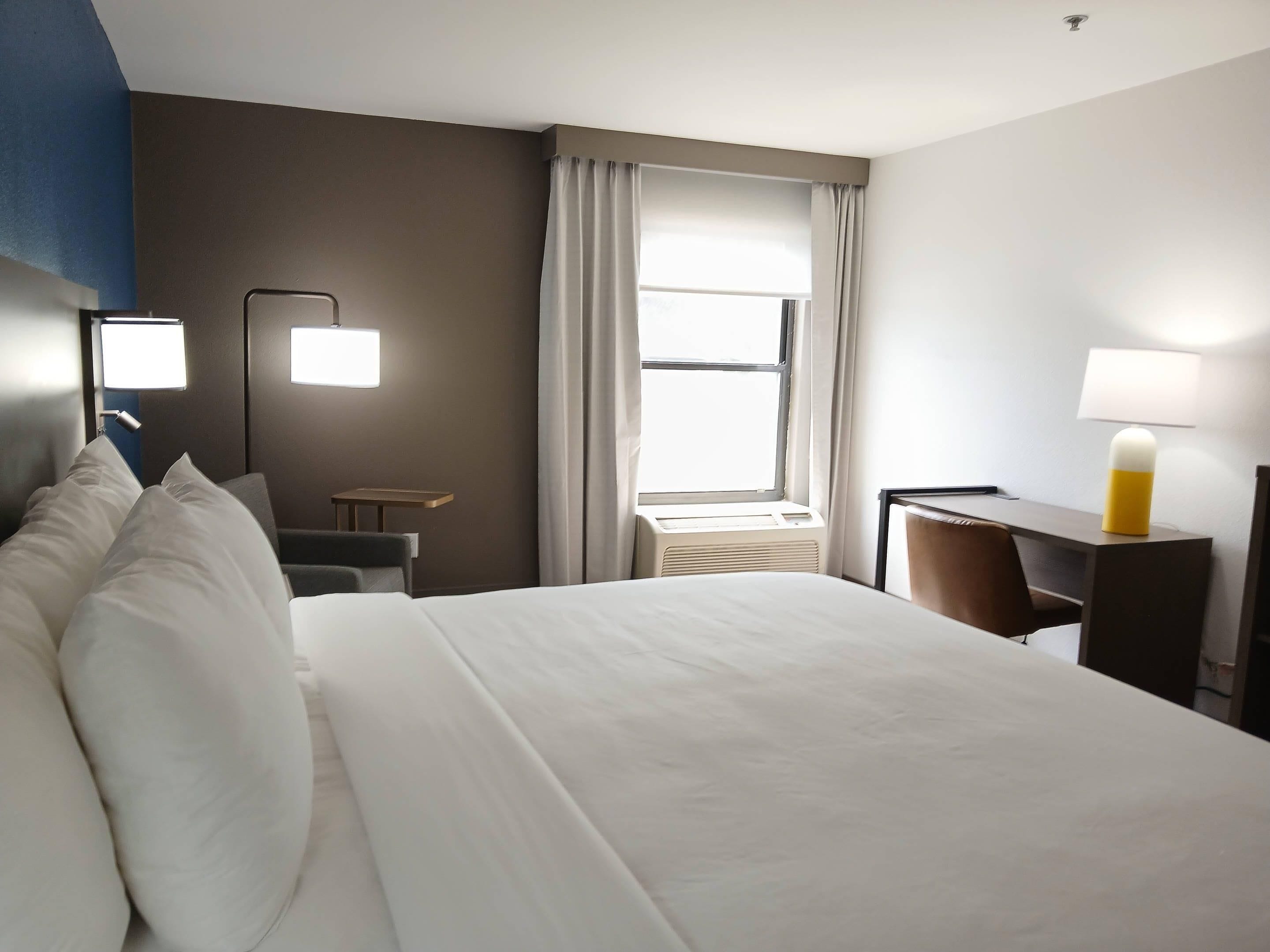 Habitación de hotel con cama, escritorio, silla, lámpara, ventana con cortinas y aire acondicionado instalado en la pared.