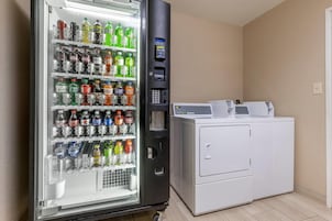 Una máquina expendedora llena de diversas bebidas y una lavadora blanca en una habitación con paredes color crema.