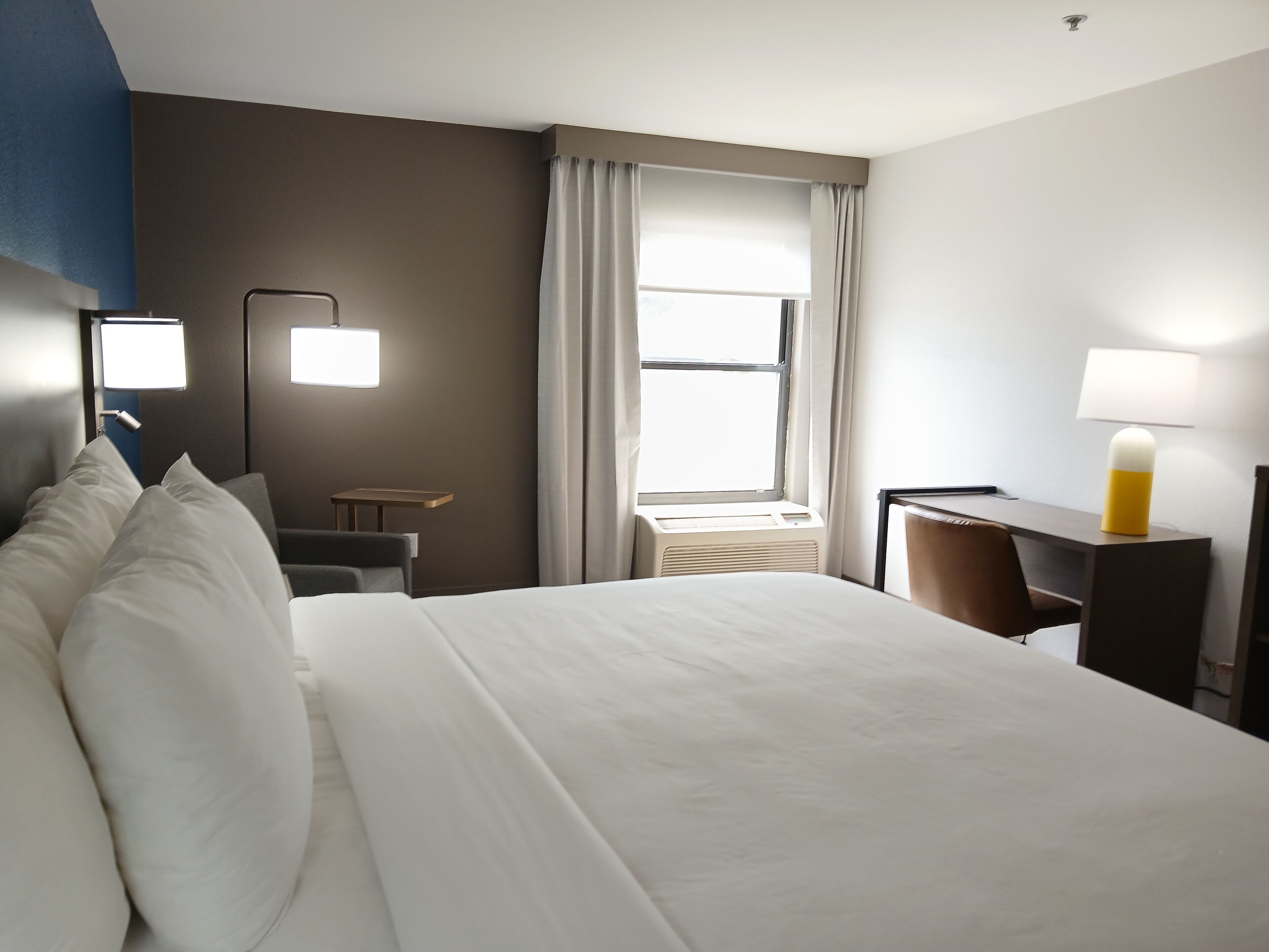 A hotel room with a large bed, a desk, a chair, a lamp, and a window with curtains.