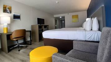 A hotel room with a bed, a desk with a chair, a TV, a yellow ottoman, and artwork on the walls.