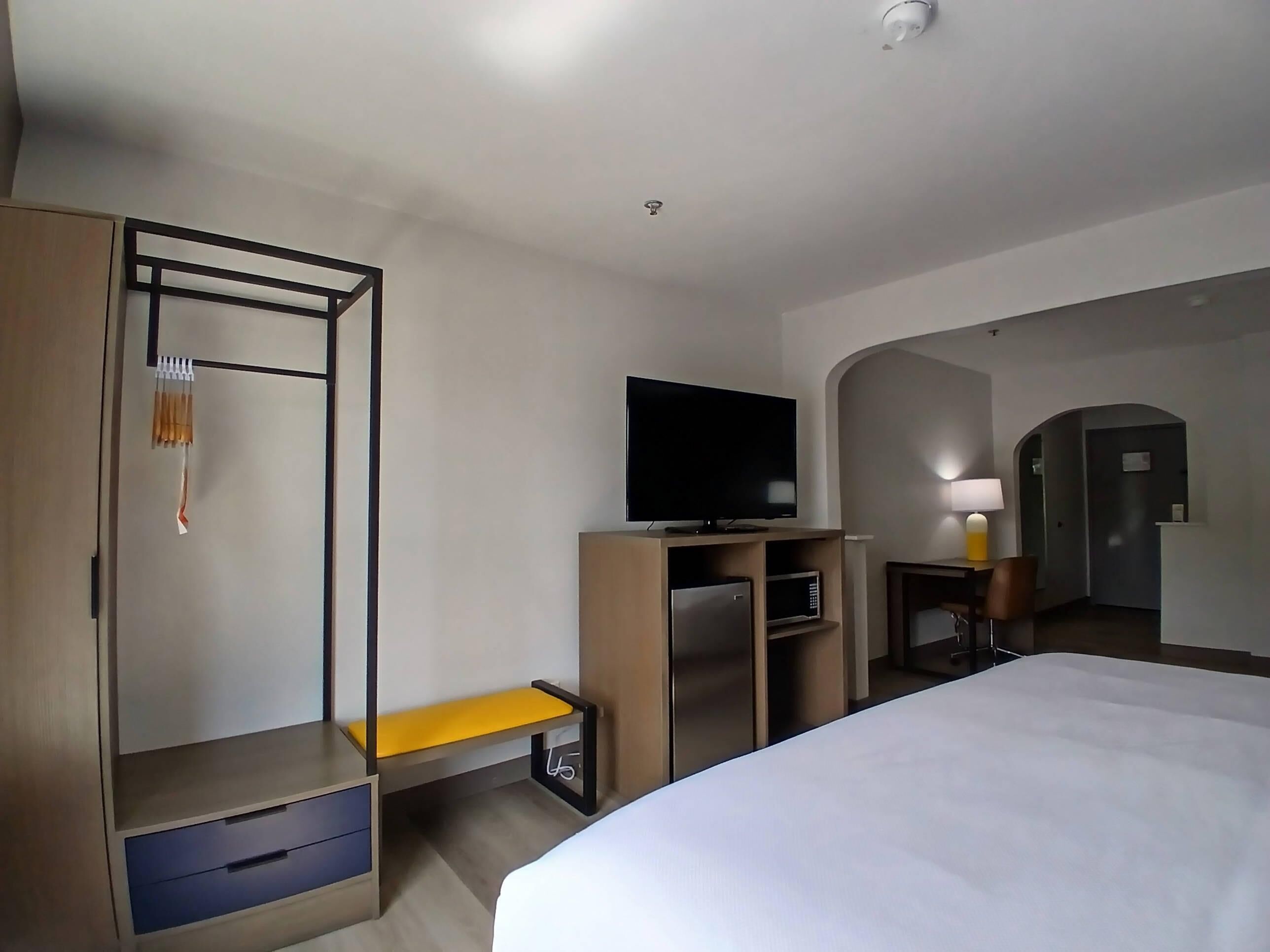 Una habitación de hotel con cama, televisor, escritorio con silla, armario y un banco.