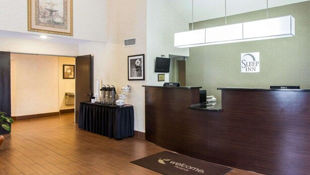 A hotel reception area with a dark wood reception desk, a welcome mat, a framed picture on the wall, and a large window above the desk. - Sleep Inn Florence North (Florence)