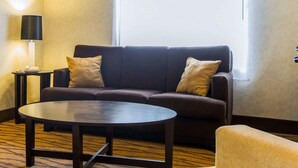 A hotel room with a dark-colored sofa, two pillows, a small round table, a lamp, and a large window with blinds. - Sleep Inn Florence North (Florence)