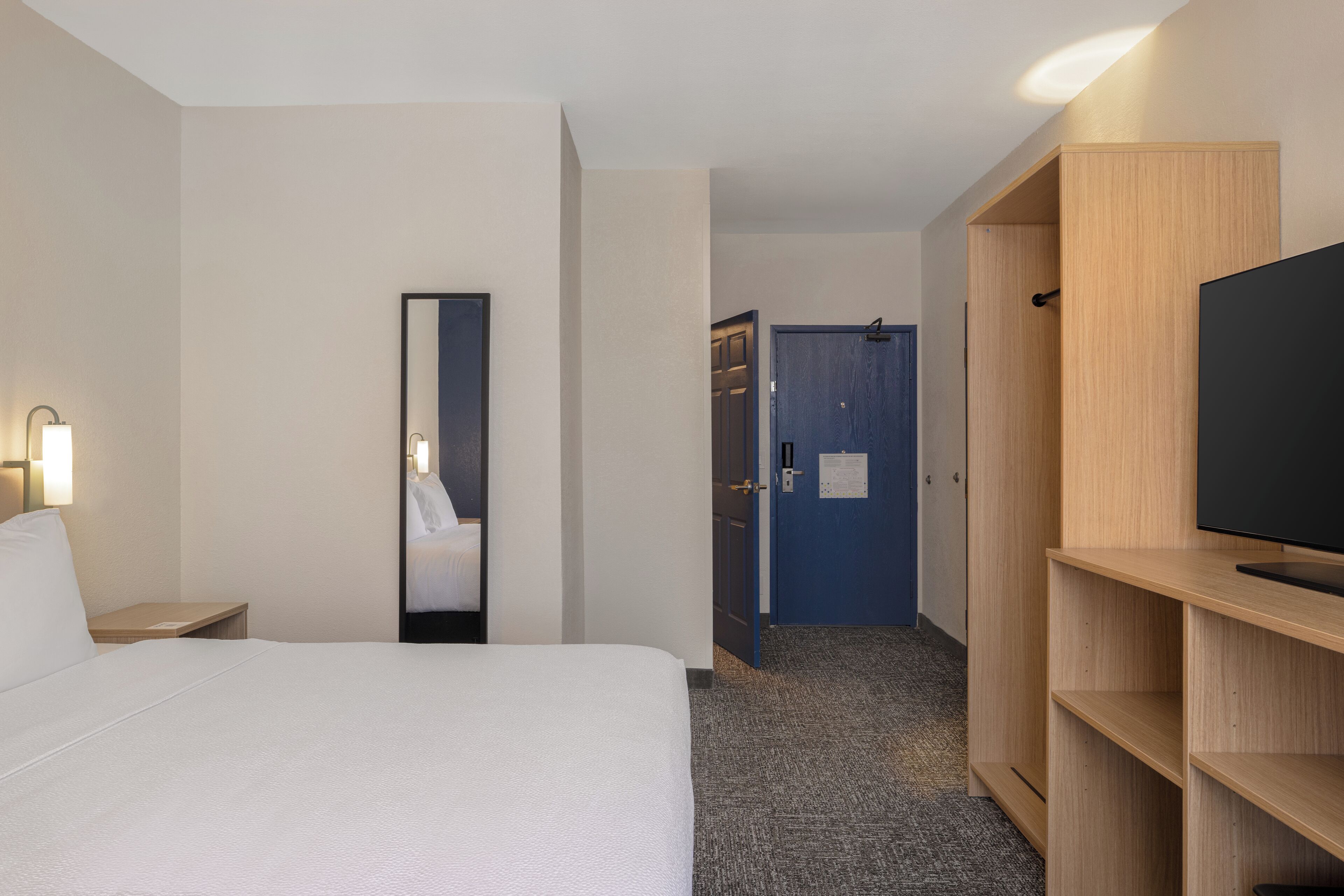 Een hotelkamer met een bed, een televisie op een houten standaard, een spiegel, een deur en een wandlamp.