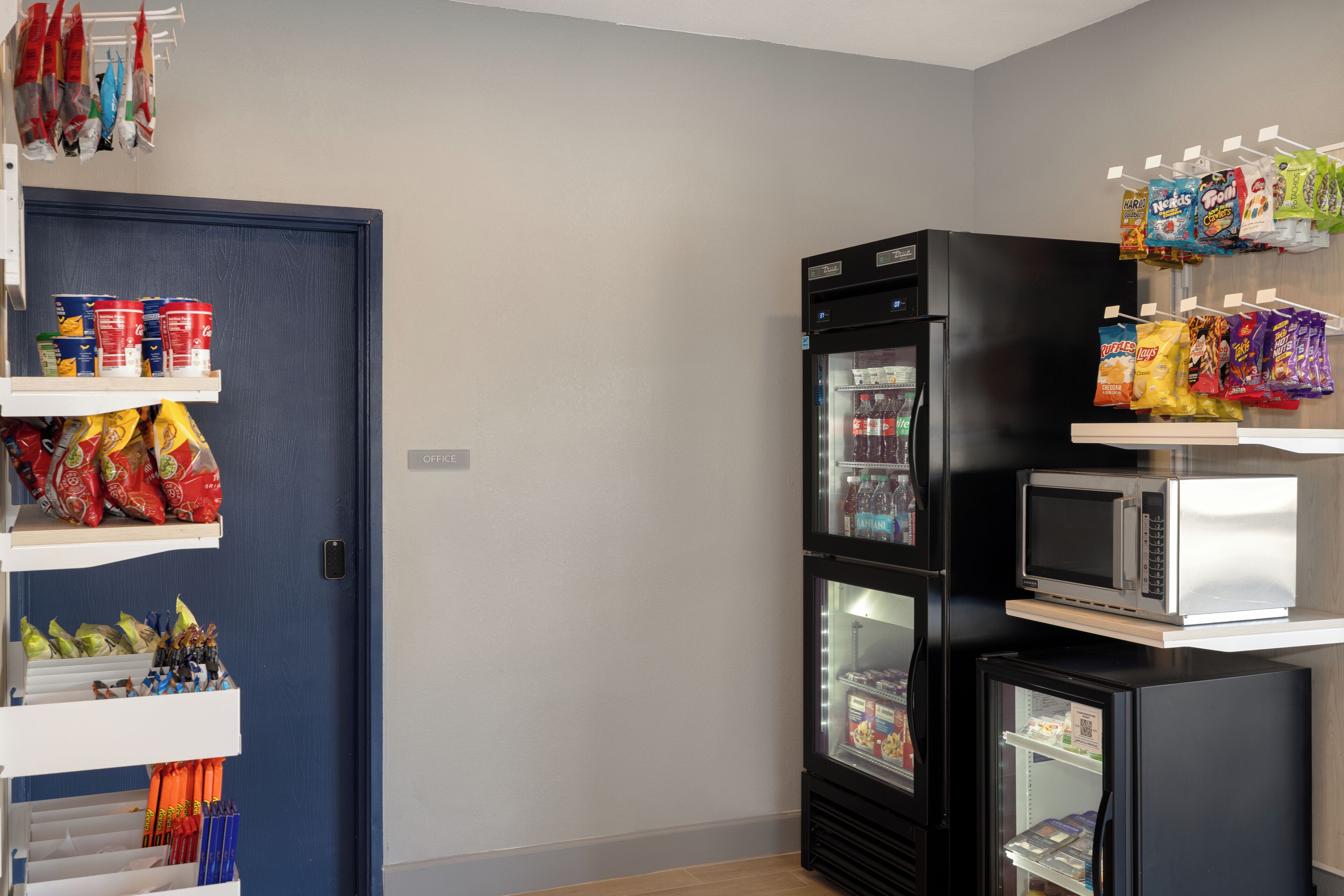 Une petite cuisine équipée d’un réfrigérateur, d’un four à micro-ondes et d’étagères remplies de collations et de boissons.