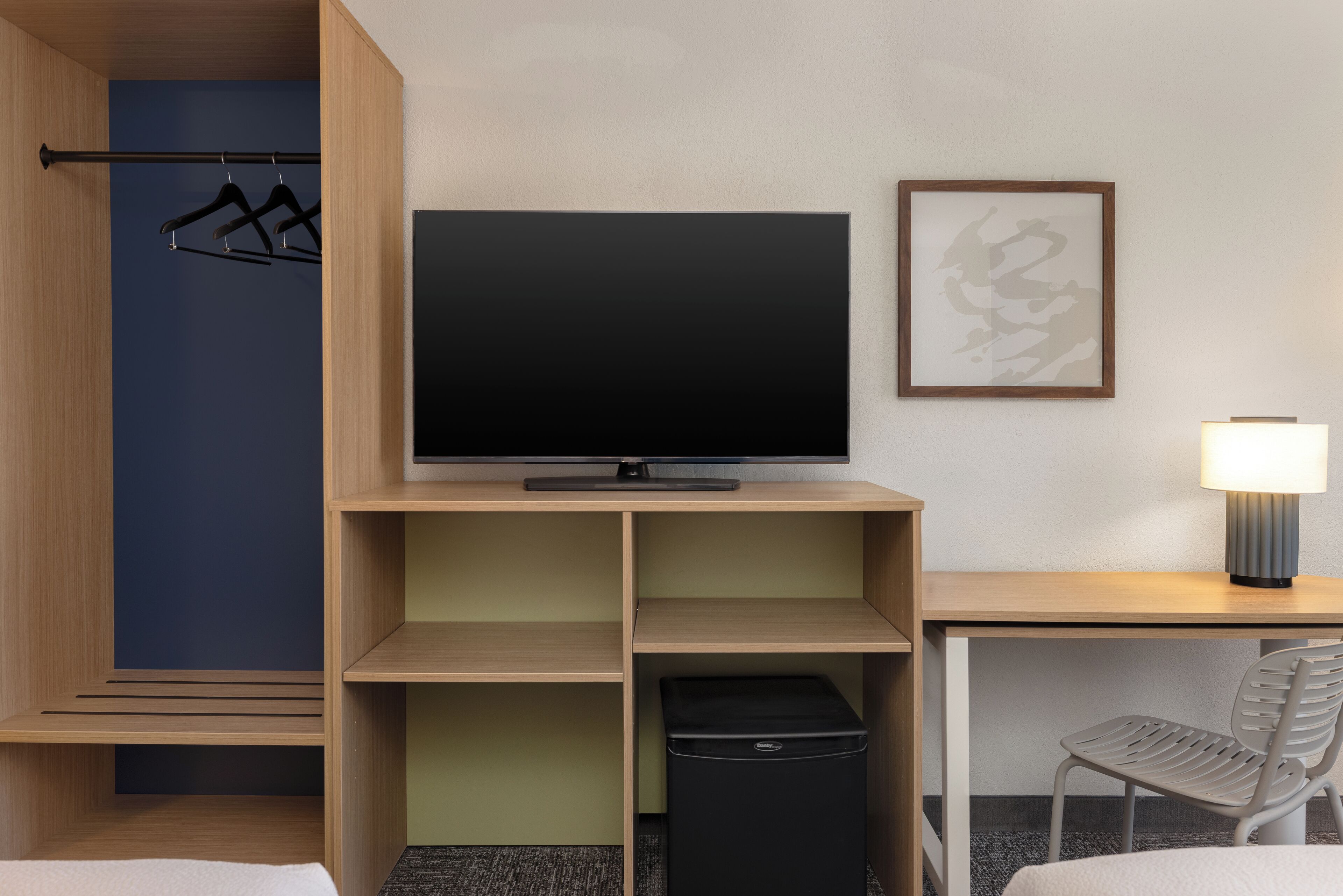 Una habitación de hotel con una cama, un escritorio con lámpara, un televisor, un cubo de basura y un perchero.
