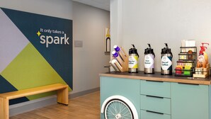 A coffee station with a wall sign saying "It only takes a spark," a wooden bench, and a teal-colored cabinet with drawers. - Spark by Hilton El Paso University (El Paso)