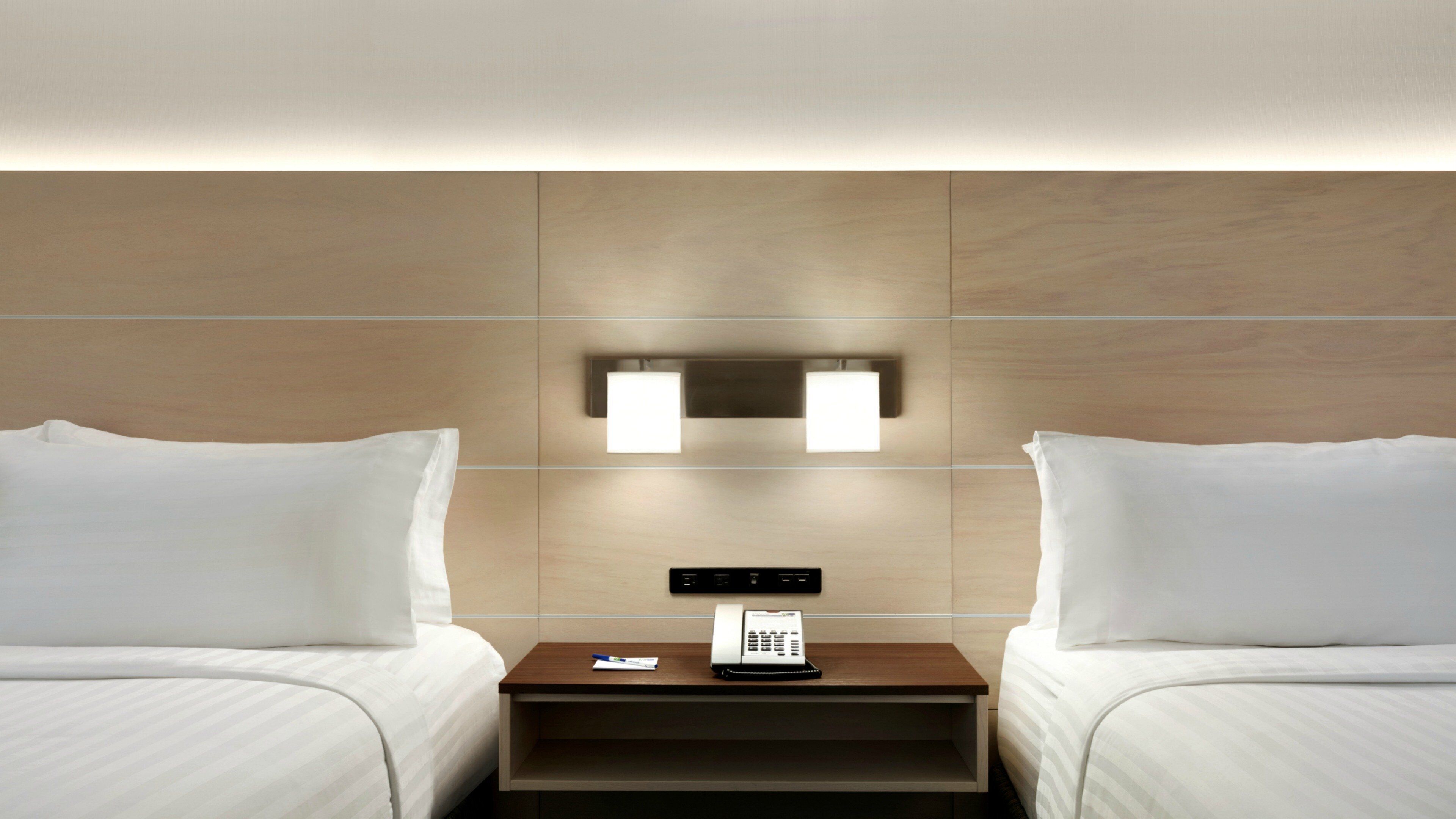 A hotel room with two beds, a nightstand with a phone, and wall-mounted lighting.