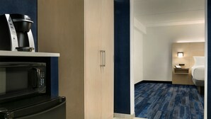 A hotel room with a kitchenette including a coffee maker, microwave, and oven. There is a bed with a nightstand, a lamp, and a small table. The room has a blue carpet and wooden walls. - Holiday Inn Express Williamsbrg Busch Gardens Area by IHG (Williamsburg)