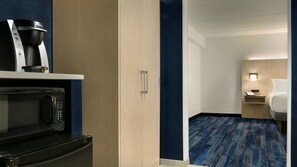 A hotel room with a kitchenette including a coffee maker, microwave, and oven. There is a bed with a nightstand, a lamp, and a small table. The room has a blue carpet and wooden walls. - Holiday Inn Express Williamsbrg Busch Gardens Area by IHG (Williamsburg)