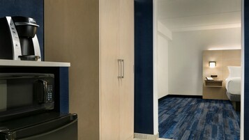 Habitación de hotel con kitchenette que incluye cafetera, microondas y horno. Tiene una cama con mesita de noche, lámpara y una pequeña mesa. La habitación tiene alfombra azul y paredes de madera.