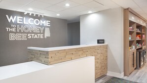 Ein Empfangsbereich mit einem Schild, auf dem 'Welcome to the Honey Bee State' steht, einem Empfangstresen, einem Hilton-Wappen und einem Schrank mit Regalen.
