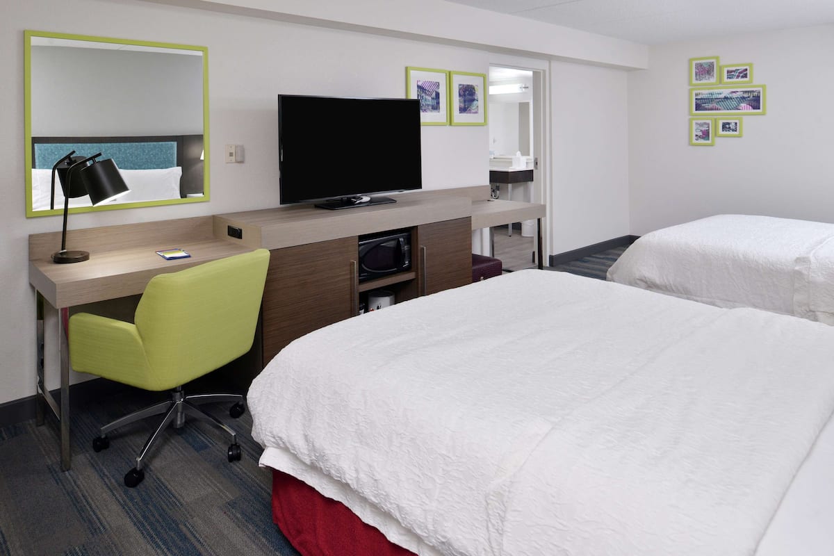 Une chambre d’hôtel équipée d’un bureau, d’une télévision, d’un fauteuil vert et de deux lits.