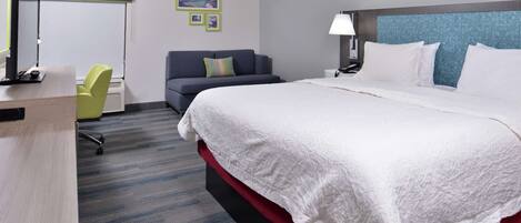 A hotel room with a bed, a desk, a chair, a lamp, and a wall with framed pictures.