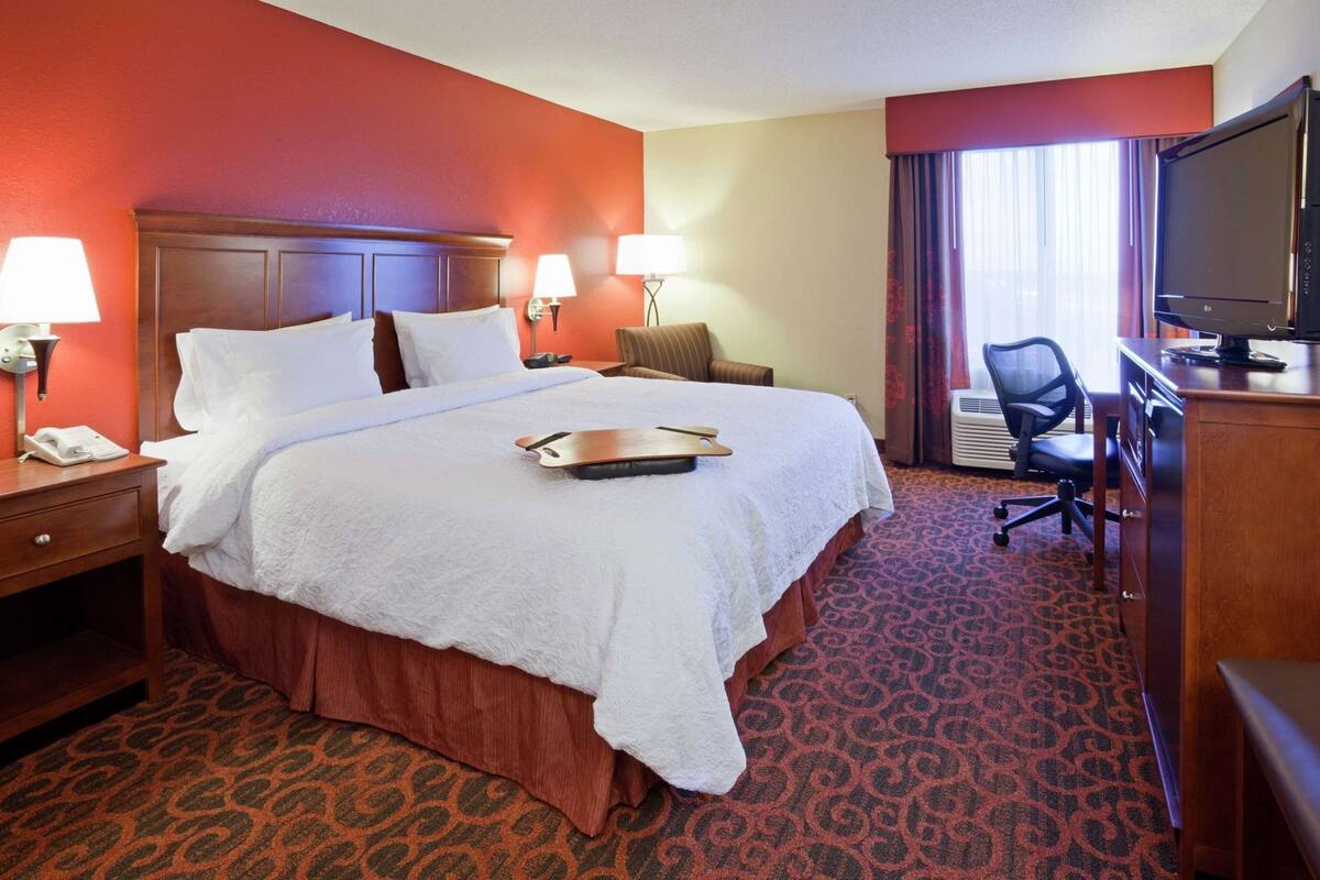 A hotel room with a large bed, two bedside lamps, a desk with a TV, a chair, and a patterned carpet.