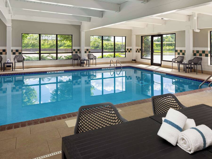 Una piscina con sillones de descanso y una mesa, amplias ventanas con vistas a zonas verdes y suelo de baldosas.