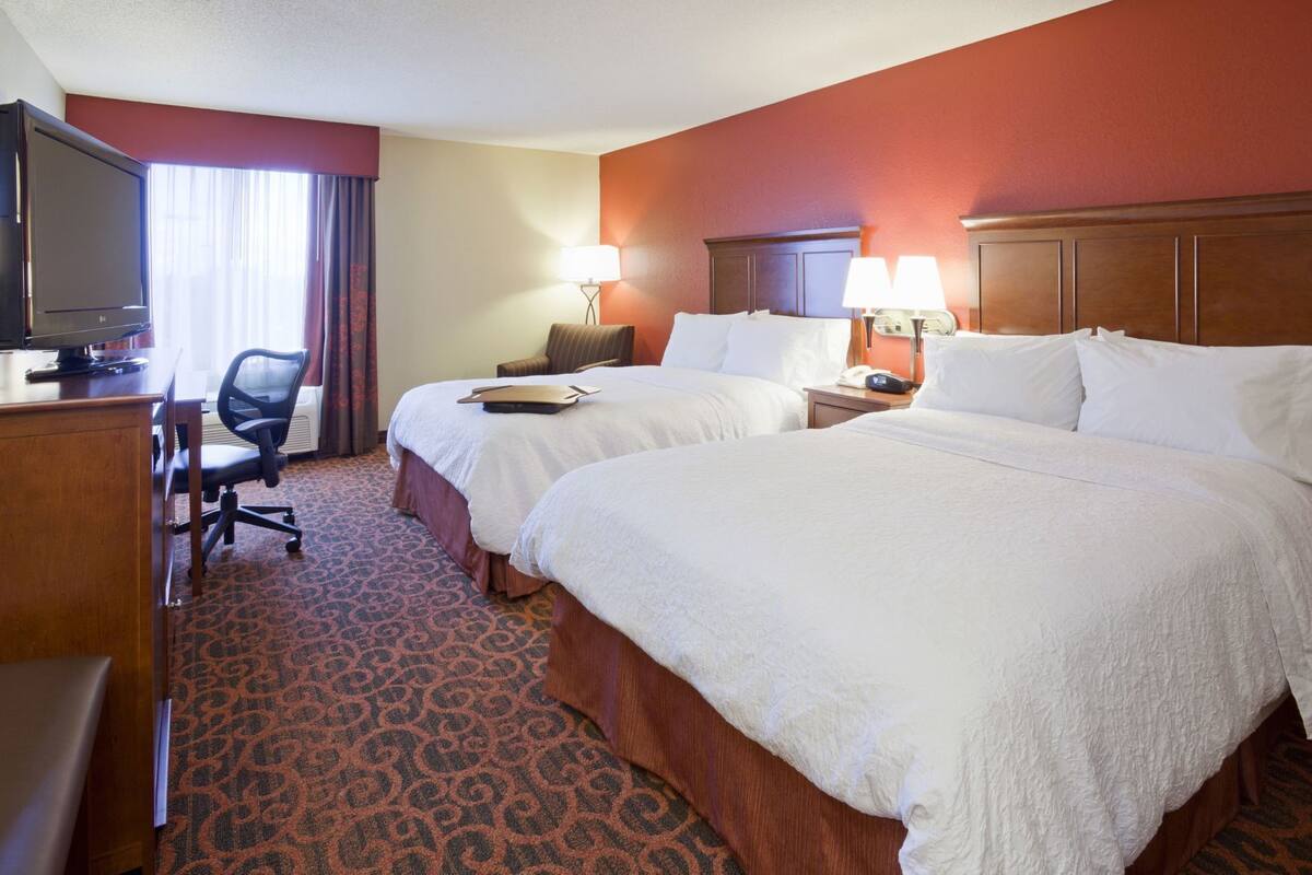 A hotel room with two beds, a desk with a TV, a chair, and a lamp.