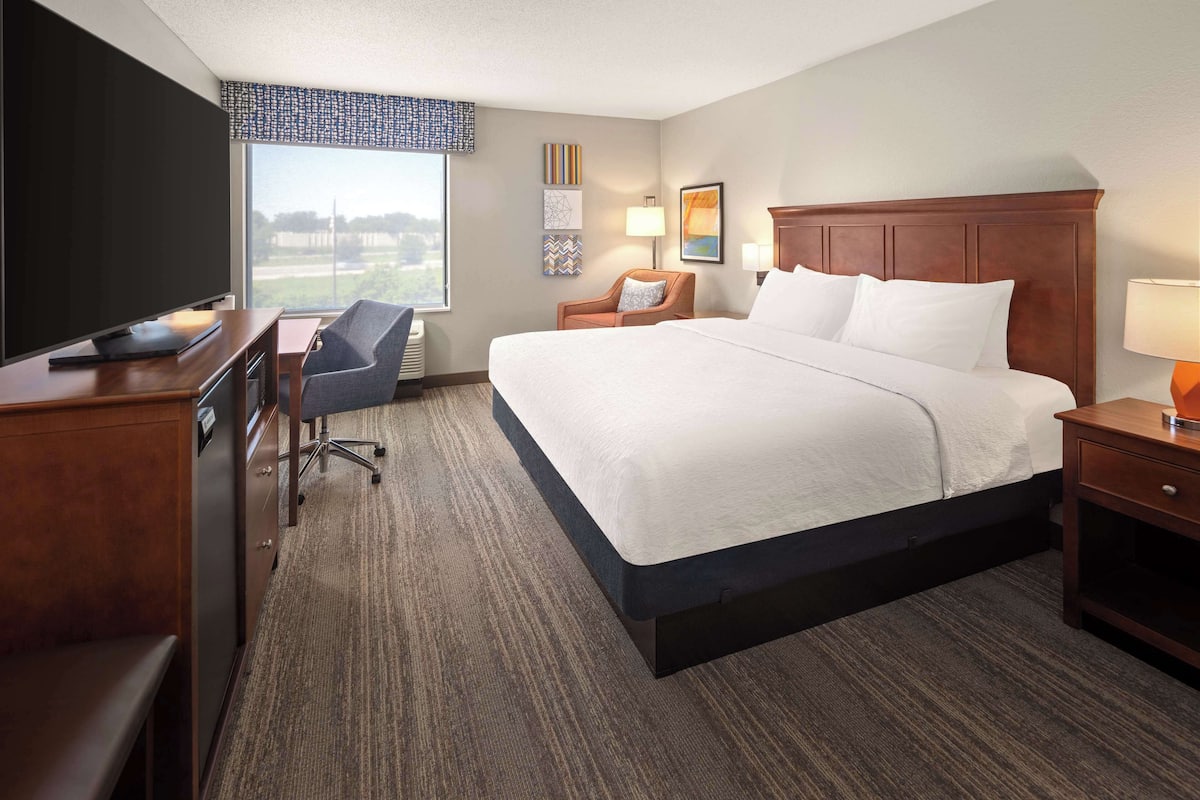 A hotel room with a large bed, a desk with a flat-screen TV, a chair, a nightstand with a lamp, and a window with a view.