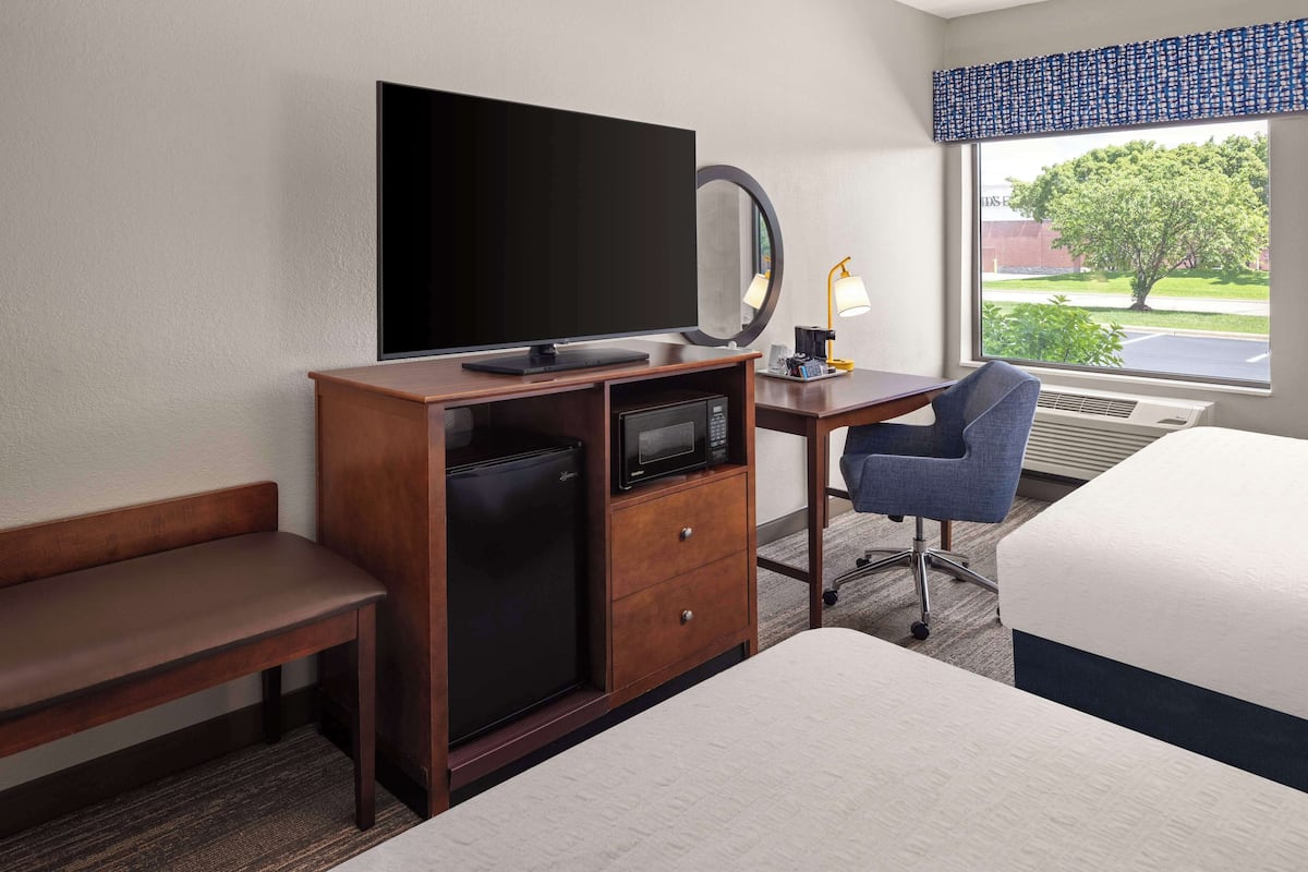 A hotel room with a bed, a desk with a computer, a small refrigerator, a TV, a mirror, and a window with a view of trees and a building.