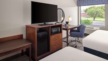 A hotel room with a bed, a desk with a computer, a small refrigerator, a TV, a mirror, and a window with a view of trees and a building.