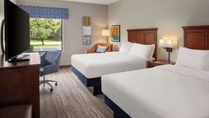 A hotel room with two beds, a desk with a TV, a chair, and a window with a view of trees.
