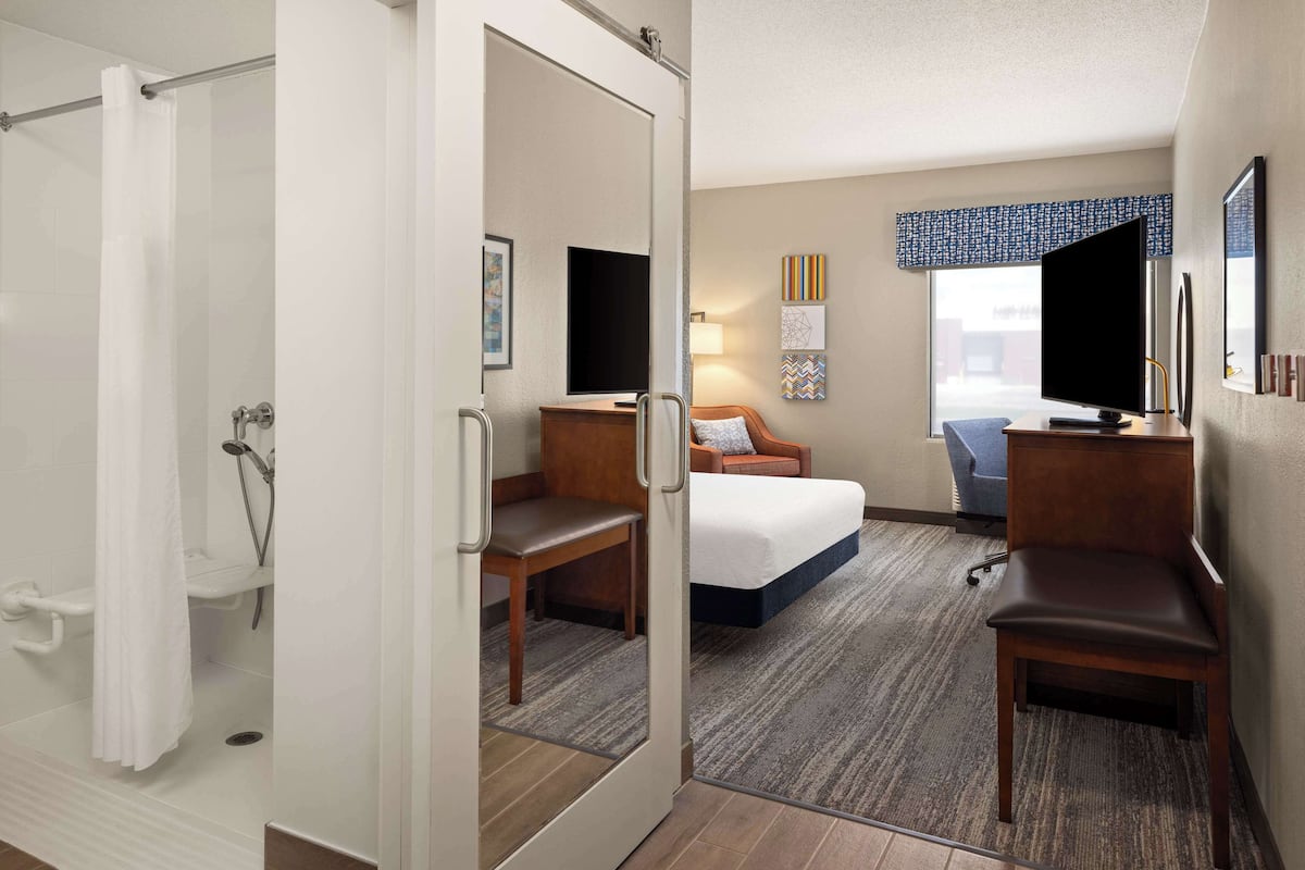 A hotel room with a shower, a bed, a desk, a chair, and a TV.