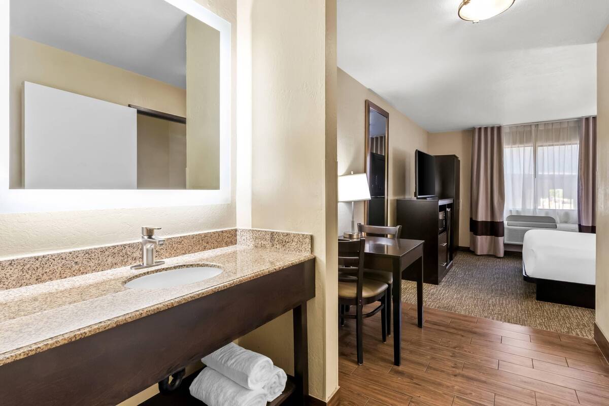 Habitación de hotel con un espejo grande, un lavamanos, un escritorio con silla, un televisor y una cama.