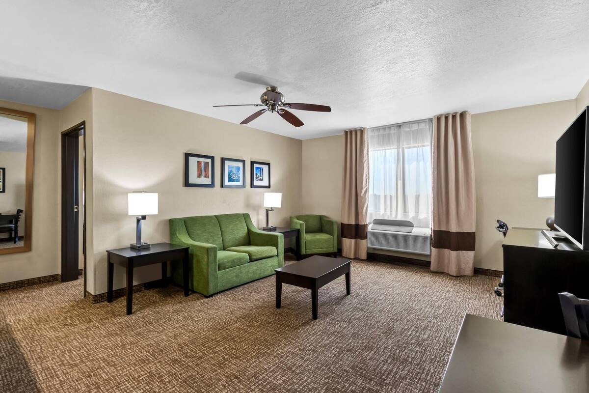 Habitación de hotel con sofá verde, una mesita, ventilador de techo y ventana con cortinas.