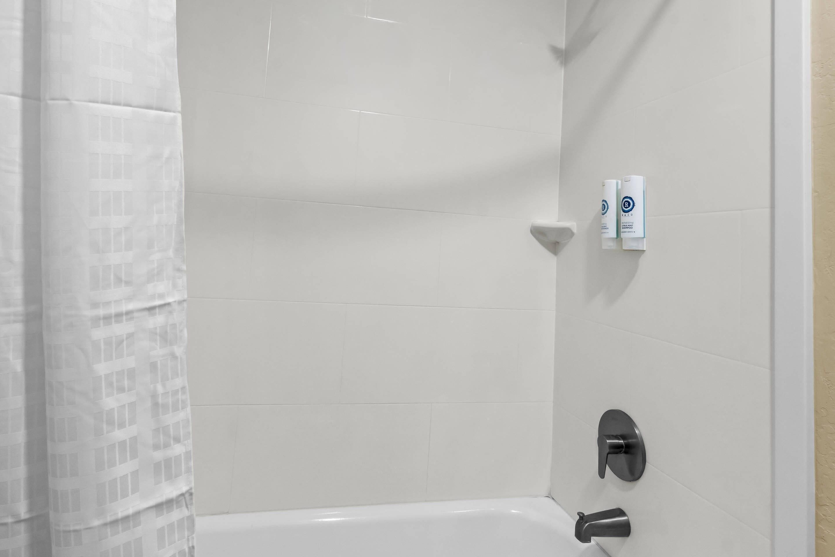 A white tiled bathroom with a shower curtain, a bathtub, and two hand dryers mounted on the wall.